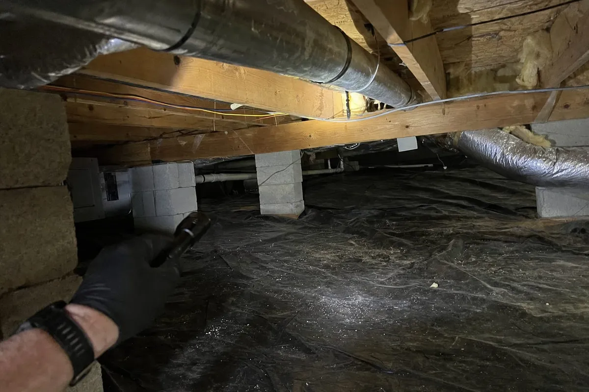 Inside a dark crawlspace, an inspector's gloved hand holds a flashlight over the vapor barrier